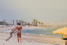 A person in a bikini stands on a sandy beach, holding a colorful towel that is caught by the wind. Nearby, an orange beach umbrella partially covers some beach gear. The ocean waves lap against the shore, and palm trees are visible in the background under a hazy sky.