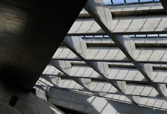 A geometric architectural structure with a grid-like ceiling composed of concrete beams casting shadows on the surfaces beneath. The ceiling allows natural light to filter through, illuminating the space. There are metal railings and a lamp post visible, suggesting this might be part of a building or public structure.