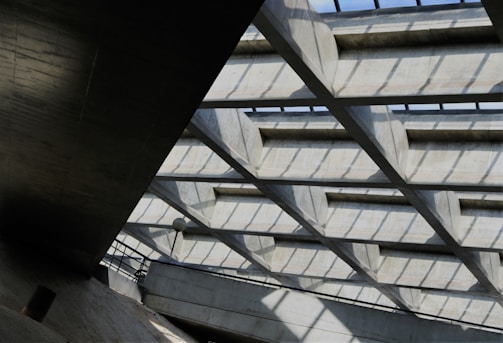 A geometric architectural structure with a grid-like ceiling composed of concrete beams casting shadows on the surfaces beneath. The ceiling allows natural light to filter through, illuminating the space. There are metal railings and a lamp post visible, suggesting this might be part of a building or public structure.