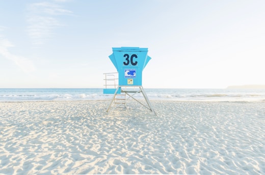 A bright and tranquil beach scene with a turquoise lifeguard tower marked '3C' standing on the sand. Gentle ocean waves roll onto the shore under a clear, pale blue sky, creating a peaceful and calm atmosphere.