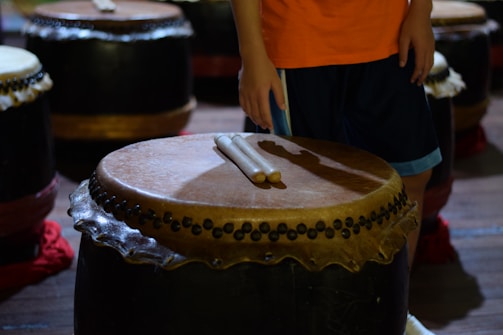 A person is standing next to a large traditional drum with drumsticks placed on top. The drum features a decorative rim and is surrounded by several similar drums. The person is wearing an orange shirt and dark shorts.