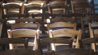Wooden chairs arranged in rows, with sunlight filtering through and highlighting certain sections. The chairs are identical and have the letters 'SEP' carved into the backrest. The lighting creates a play of shadows and light, adding depth to the scene.