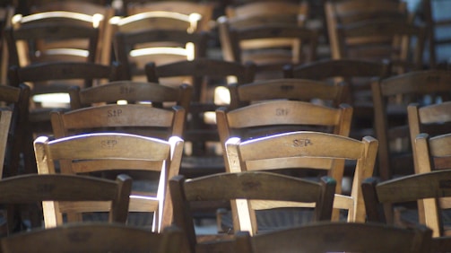 Wooden chairs arranged in rows, with sunlight filtering through and highlighting certain sections. The chairs are identical and have the letters 'SEP' carved into the backrest. The lighting creates a play of shadows and light, adding depth to the scene.