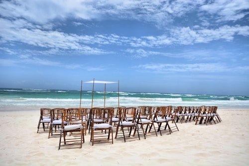 A serene beach setting with rows of wooden chairs arranged on the sandy shore, facing the ocean. The sky is partly cloudy, and the turquoise waves roll gently in the background. A simple, minimalistic canopy structure is set up, suggesting a possible beach event or wedding.