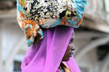 A person is wearing a vibrant headscarf with a colorful and intricate pattern, combined with a sheer, bright purple veil. The background is softly blurred, suggesting outdoor surroundings.