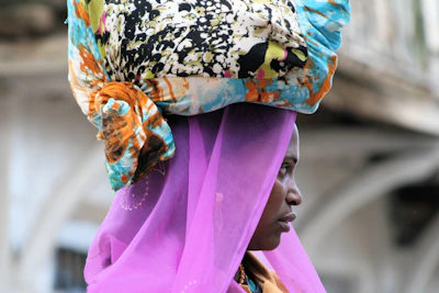 A person is wearing a vibrant headscarf with a colorful and intricate pattern, combined with a sheer, bright purple veil. The background is softly blurred, suggesting outdoor surroundings.