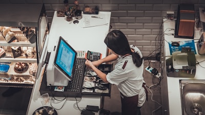 A person is organizing a cash register in a cafe or bakery setting. The counter is equipped with a computer, card reader, and cash drawer. On the left, there is a display case filled with various cakes and pastries. The workspace is surrounded by various tools and equipment, indicating a busy and organized environment.
