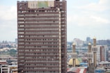 A tall, rectangular high-rise building with many windows is situated in an urban environment. The cityscape includes numerous buildings in the background and several mosque minarets with golden domes nearby. The sky is partially cloudy, contributing to a neutral lighting effect.