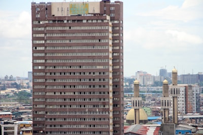 A tall, rectangular high-rise building with many windows is situated in an urban environment. The cityscape includes numerous buildings in the background and several mosque minarets with golden domes nearby. The sky is partially cloudy, contributing to a neutral lighting effect.