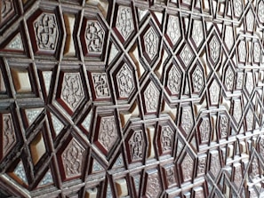 A detailed geometric pattern featuring a series of interlocking, raised hexagonal and diamond shapes. Each shape contains ornate engraved designs, resembling traditional or historical motifs. The texture appears to be carved wood, giving a sense of depth and craftsmanship.