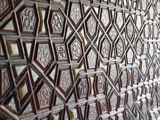 A detailed geometric pattern featuring a series of interlocking, raised hexagonal and diamond shapes. Each shape contains ornate engraved designs, resembling traditional or historical motifs. The texture appears to be carved wood, giving a sense of depth and craftsmanship.