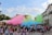 A large group of people participates in a colorful outdoor festival, with vibrant clouds of pink, green, yellow, and blue powder filling the air. The crowd, mostly wearing white T-shirts, is gathered in a paved public space surrounded by buildings and greenery. Some participants have raised their arms, and many are covered in colorful powder.