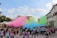 A large group of people participates in a colorful outdoor festival, with vibrant clouds of pink, green, yellow, and blue powder filling the air. The crowd, mostly wearing white T-shirts, is gathered in a paved public space surrounded by buildings and greenery. Some participants have raised their arms, and many are covered in colorful powder.