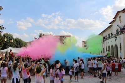 A large group of people participates in a colorful outdoor festival, with vibrant clouds of pink, green, yellow, and blue powder filling the air. The crowd, mostly wearing white T-shirts, is gathered in a paved public space surrounded by buildings and greenery. Some participants have raised their arms, and many are covered in colorful powder.