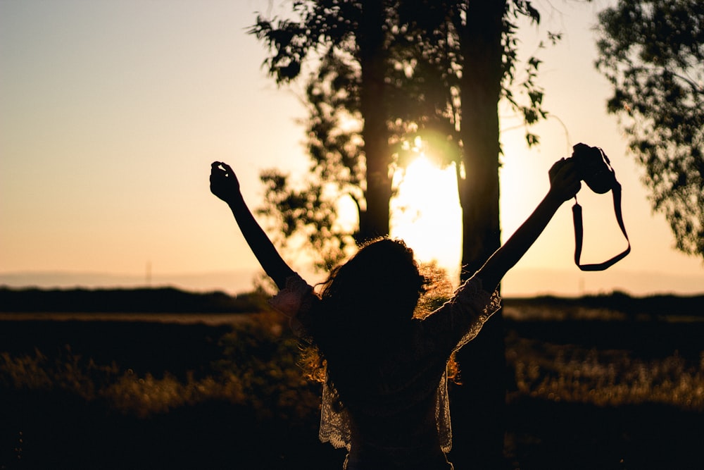A woman stretches her arms in the air, holding a camera as a golden sunset illuminates the sky