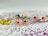A collection of colorful stud earrings is displayed on a surface with scattered purple glitter. Each earring set has a distinct design, including smiley faces, multicolored petals, and flowers with red or pink centers.