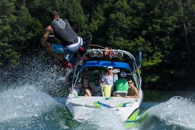 A person is performing a jump on a wakeboard, pulled by a motorboat across a body of water. The boat is occupied by several people, and water splashes around them. Tall trees are visible in the background, indicating a natural, outdoor setting.