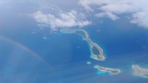 An aerial view of a small chain of islands surrounded by clear blue ocean waters. The islands are outlined with white sandy beaches and dark green vegetation. A subtle rainbow arcs across the sky, partially obscured by light clouds.