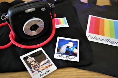 A black instant camera with a red strap is placed on a black surface. Nearby, there are two Polaroid photos; one depicts a famous London landmark and the other shows a person interacting with a dolphin. There are colorful labels with the text 'PolaroMagnet' in the scene.