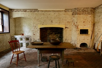 A rustic, old-fashioned kitchen with exposed brick walls and a large, dark fireplace. The room is sparsely furnished with a wooden table and several stools, along with a red wooden chair. Various kitchen utensils and a broom are present, contributing to the vintage atmosphere. The lighting is dim, originating from a window on the left, casting gentle shadows around the room.