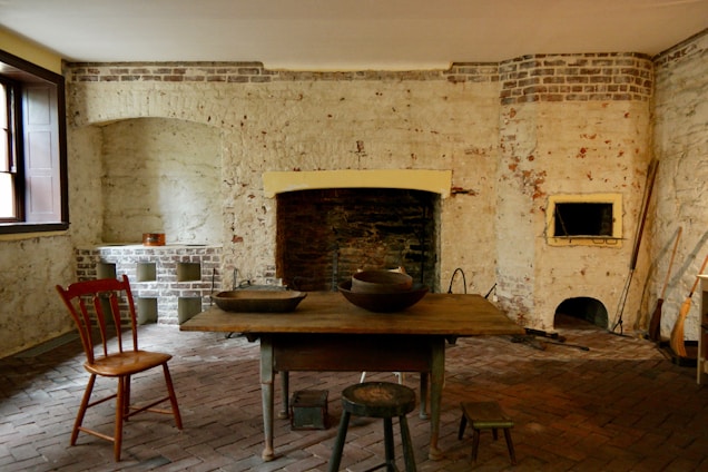 A rustic, old-fashioned kitchen with exposed brick walls and a large, dark fireplace. The room is sparsely furnished with a wooden table and several stools, along with a red wooden chair. Various kitchen utensils and a broom are present, contributing to the vintage atmosphere. The lighting is dim, originating from a window on the left, casting gentle shadows around the room.