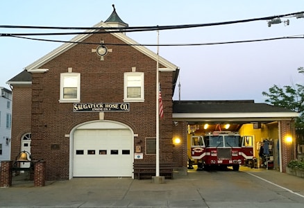 A brick fire station with an American flag, labeled 'Saugatuck Hose Co. Engine Co. 4' with a large garage housing a red fire truck. The firehouse has two upper windows and a round window at the peak, with exterior lighting on either side of the garage entrance.