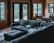 A cozy living room with wooden walls and large windows looking out onto a wooded area. A dark gray sectional sofa is in the center, with a laptop, remote controls, and some personal items spread on it. A dining area with a wooden table and chairs is visible in the background.