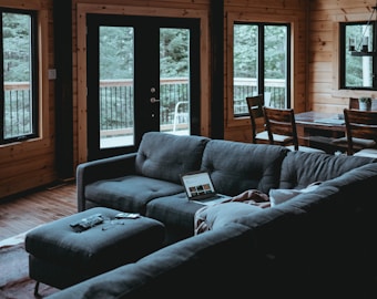 A cozy living room with wooden walls and large windows looking out onto a wooded area. A dark gray sectional sofa is in the center, with a laptop, remote controls, and some personal items spread on it. A dining area with a wooden table and chairs is visible in the background.