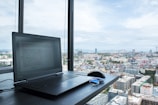 A laptop is open on a table near a window with a panoramic view of a cityscape. A computer mouse and a pair of sunglasses are placed next to the laptop. The scene captures a mix of modern technology and urban life under a partly cloudy sky.