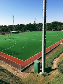 A hockey field with artificial green turf and a red boundary line is visible. Several players in sports attire are scattered across the field, indicating a game or practice session. Tall lighting poles stand around the field, and a dense area of trees is visible in the background.