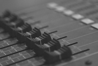 A grayscale close-up of an audio mixing console with several faders in focus, arranged in a row, likely used for adjusting sound levels during audio production.