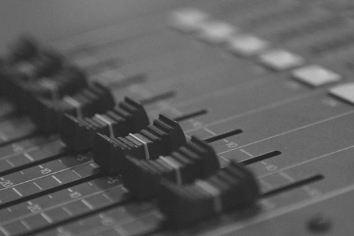 A grayscale close-up of an audio mixing console with several faders in focus, arranged in a row, likely used for adjusting sound levels during audio production.