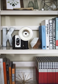A stylish bookshelf display features an antique camera with a vintage flash, assorted books, and decorative items including a metal letter, framed artwork, geometric objects, and a starburst sculpture. The arrangement combines elements of mid-century and contemporary design with emphasis on aesthetics and functionality.