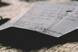 A close-up view of the corner of a handwritten letter or postcard on textured fabric. The paper shows slight creases and handwritten script in black ink, with words of affection visible. Lighting creates a warm, nostalgic ambiance.