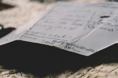 A close-up view of the corner of a handwritten letter or postcard on textured fabric. The paper shows slight creases and handwritten script in black ink, with words of affection visible. Lighting creates a warm, nostalgic ambiance.