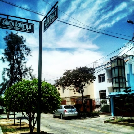 A residential street corner with a clear sky and a mix of clouds overhead. Two street signs labeled 'Santa Domitila' and 'Santa Rosalia' are prominently displayed on a pole. The area features a parked silver car, houses with modern architectural elements, and lush green trees lining the street.
