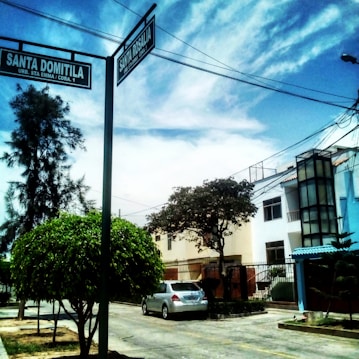A residential street corner with a clear sky and a mix of clouds overhead. Two street signs labeled 'Santa Domitila' and 'Santa Rosalia' are prominently displayed on a pole. The area features a parked silver car, houses with modern architectural elements, and lush green trees lining the street.