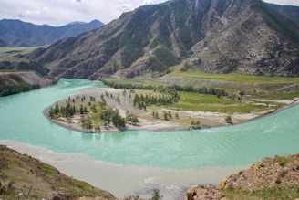 A winding turquoise river flows through a mountainous landscape with lush greenery along its banks. The river creates an island with scattered trees and patches of grass. Towering mountains in the background create a dramatic backdrop, with varying shades of green and brown across the rocky surfaces.