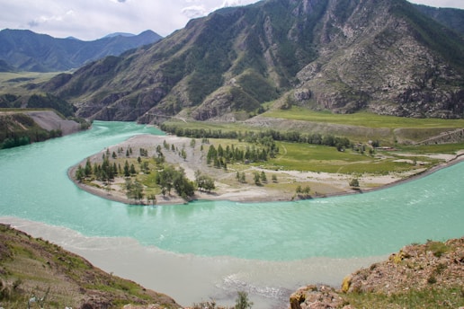 A winding turquoise river flows through a mountainous landscape with lush greenery along its banks. The river creates an island with scattered trees and patches of grass. Towering mountains in the background create a dramatic backdrop, with varying shades of green and brown across the rocky surfaces.
