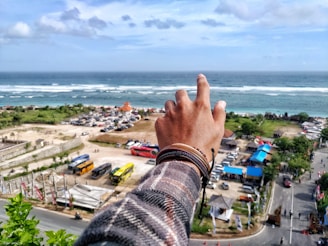 A hand wearing multiple bracelets, with fingers extended, is seen against a backdrop of a coastal area featuring the ocean, waves, and a beach. Below, there is a parking lot with several buses and vehicles, as well as a road leading through the area. There are buildings with blue roofs and green trees scattered around the landscape.