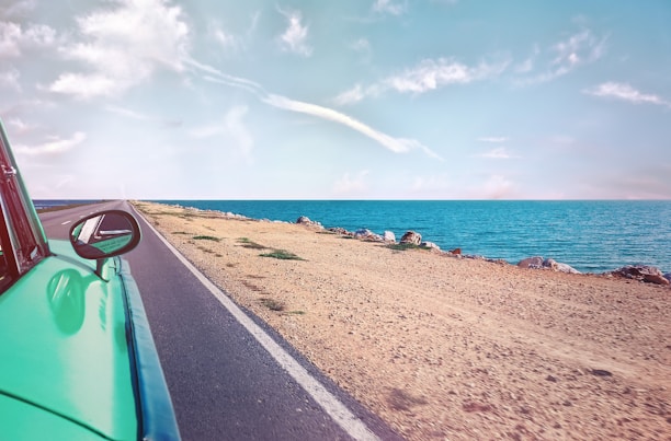 A green compact car driving along a scenic coastal road with clear blue skies.