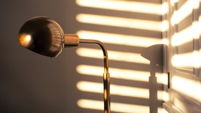 A vintage-style lamp with a brass finish is illuminated by sunlight streaming through blinds, casting horizontal shadows on the wall and creating a warm, golden ambiance around the room.
