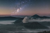 A breathtaking night scene of a mountain landscape under a starry sky with the Milky Way visible. The foreground shows a smoking volcano, surrounded by mist and silhouetted against a backdrop of other mountainous formations.