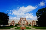 A large brick building with architectural features resembling a school or college, featuring symmetrical windows and a central entrance. A flagpole is situated in the foreground, surrounded by well-maintained lawns and trees. The sky above is bright with scattered white clouds.