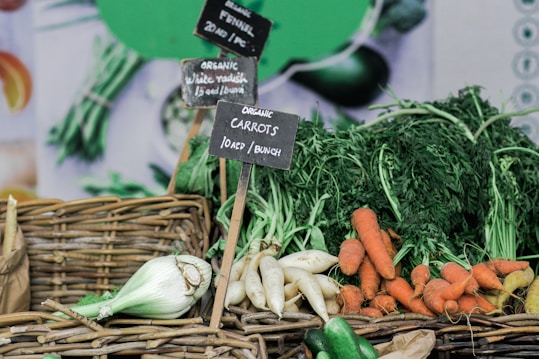A display of organic vegetables including carrots, white radishes, and fennel, arranged in woven baskets with small chalkboard signs indicating the type and price of each vegetable. Fresh produce with leafy greens adds to the vibrant and earthy appeal.