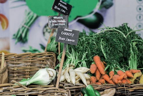 A display of organic vegetables including carrots, white radishes, and fennel, arranged in woven baskets with small chalkboard signs indicating the type and price of each vegetable. Fresh produce with leafy greens adds to the vibrant and earthy appeal.