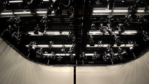 A dark studio ceiling with multiple mounted stage lights and illuminated panels in an organized grid. The surrounding walls are covered with light-colored curtains.