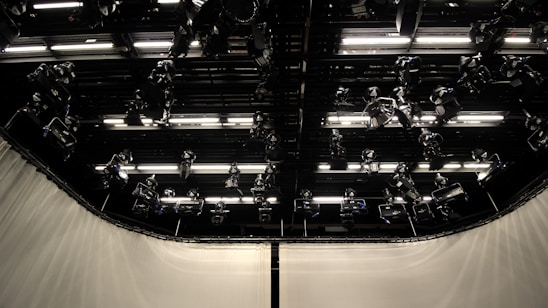 A dark studio ceiling with multiple mounted stage lights and illuminated panels in an organized grid. The surrounding walls are covered with light-colored curtains.
