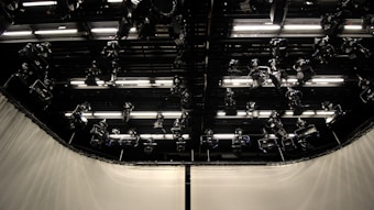 A dark studio ceiling with multiple mounted stage lights and illuminated panels in an organized grid. The surrounding walls are covered with light-colored curtains.
