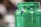 A close-up view of a green plastic jar with paint oozing from the top. The jar is prominently focused, and there are small patches of purple and yellow on the surface. The background is blurred and has a mix of brown and green tones, indicating an indoor setting.